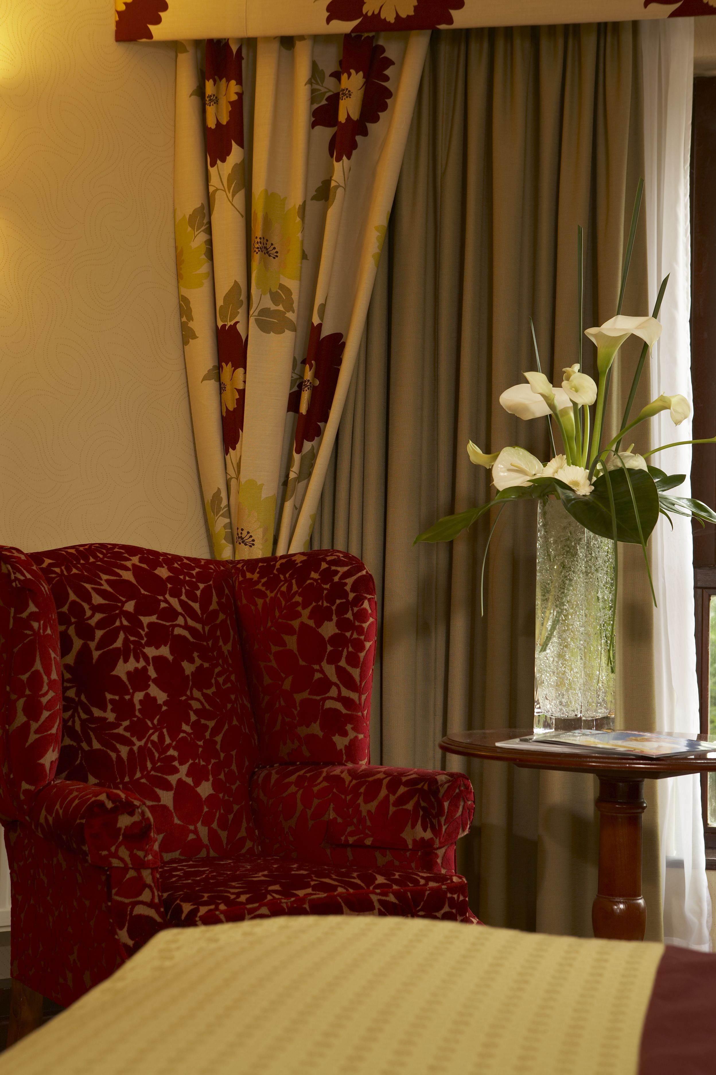 A hotel room with a red floral sofa, a small table with a vase of white flowers, and a bed with a patterned bedspread.