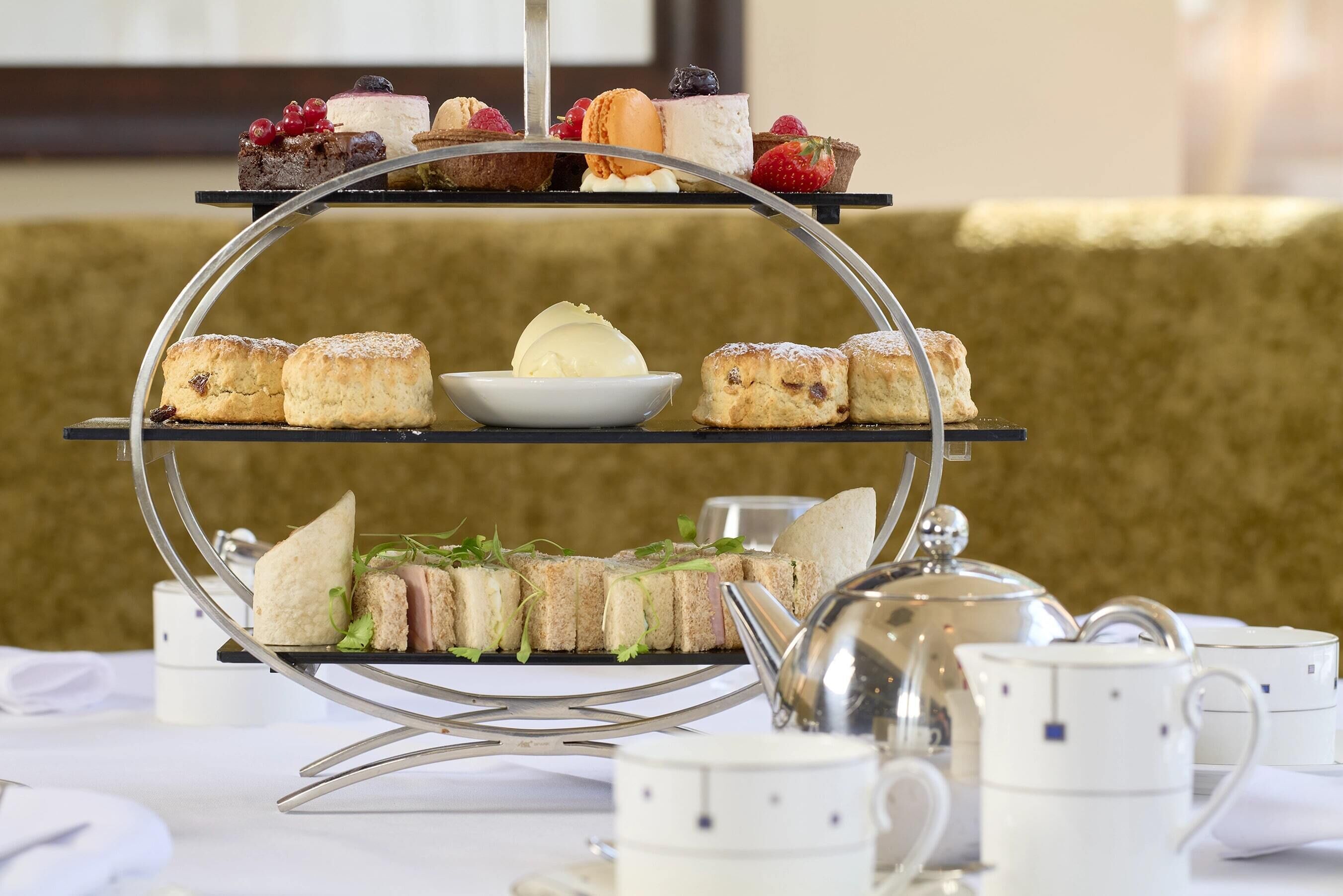 Un présentoir à trois niveaux, garni de diverses pâtisseries et surmonté d’un bol de crème glacée, accompagné de théières et de tasses, le tout disposé sur une table.