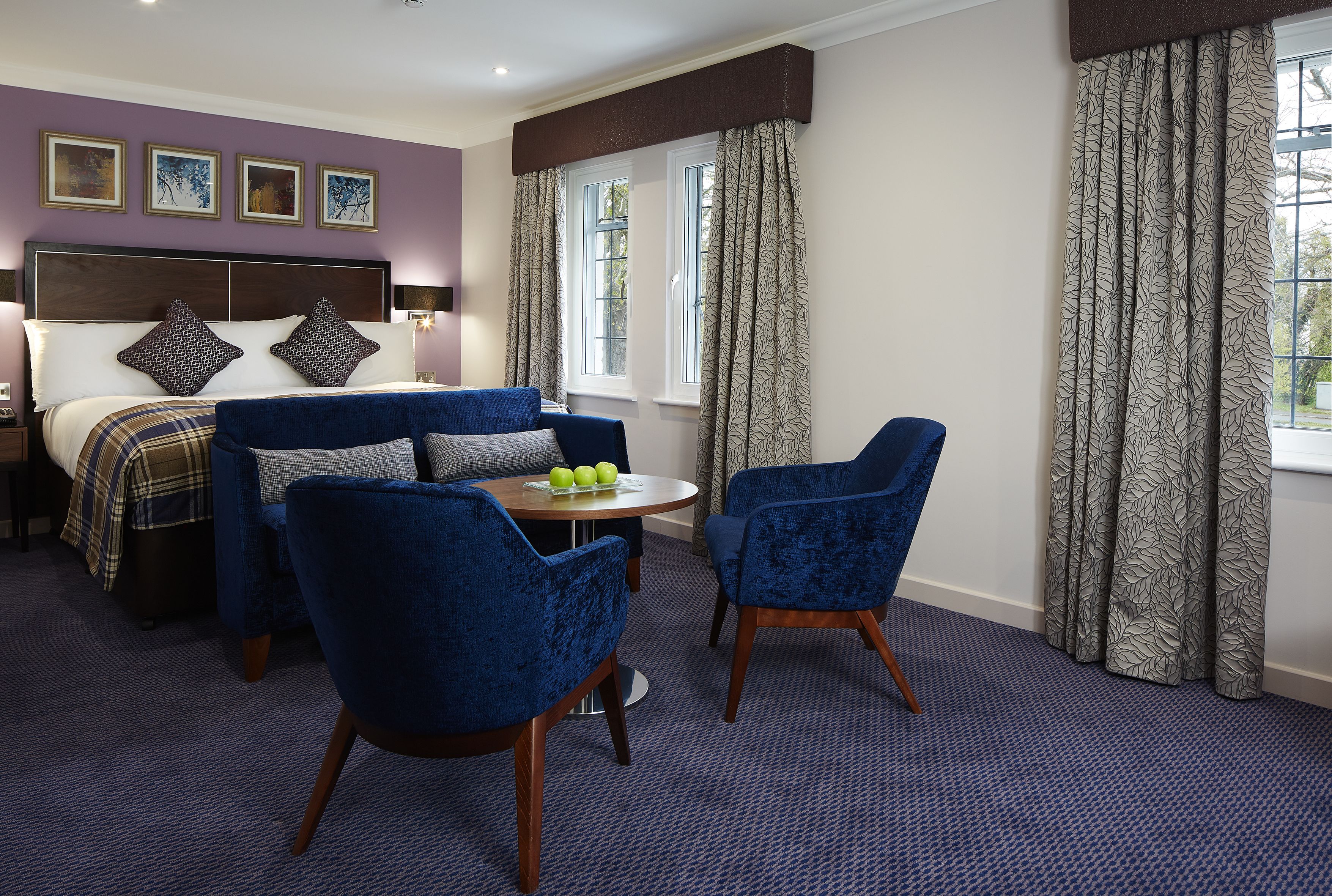 A hotel room with a bed, two blue armchairs, a small round table with apples, and a window with curtains.