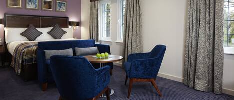 Una habitación de hotel con una cama, dos sillones azules, una mesita redonda con manzanas y un ventanal con cortinas.