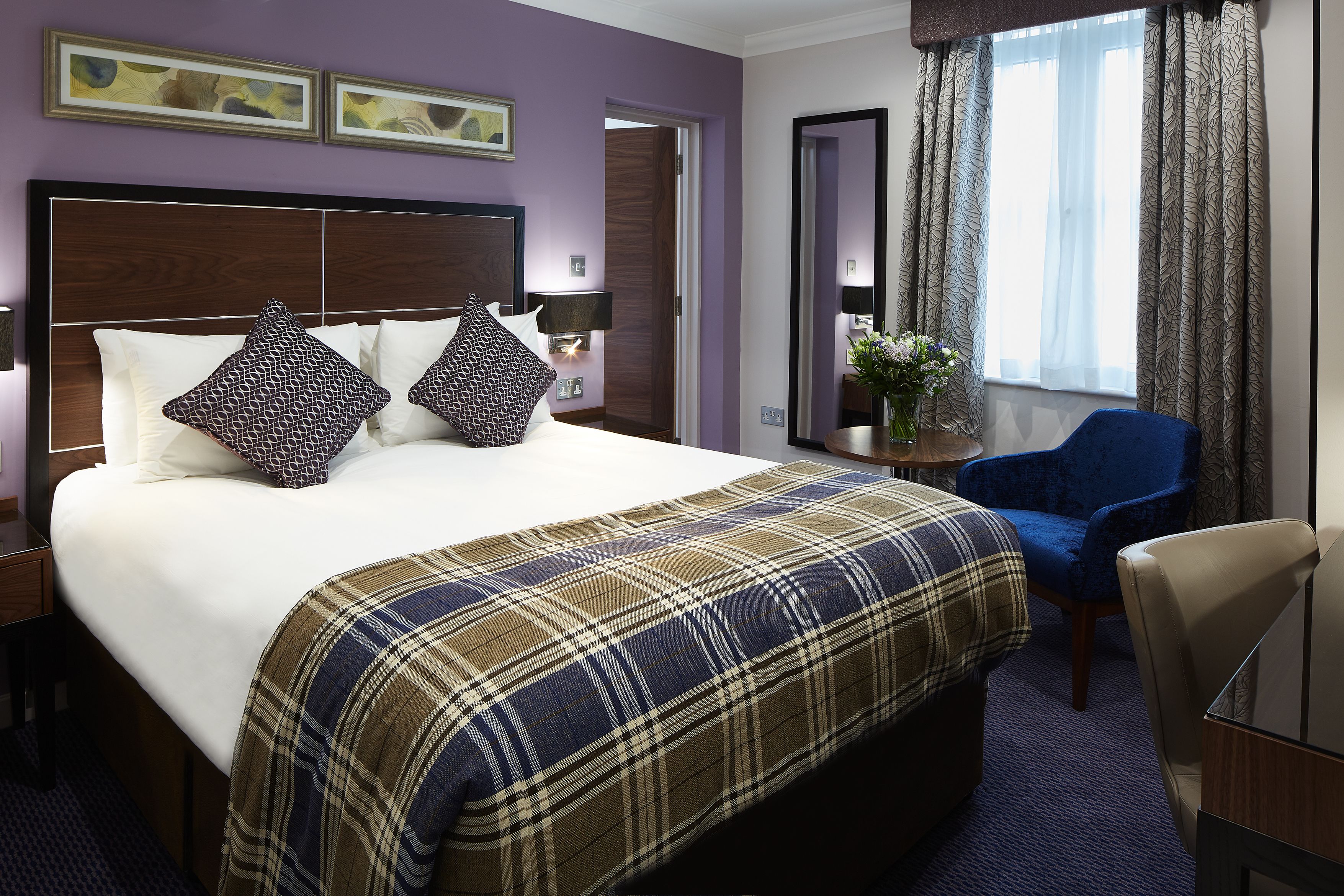 A hotel room with a large bed, a blue armchair, a wooden nightstand, and a mirror.