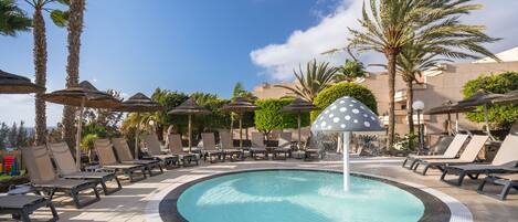 A pool area with lounge chairs and umbrellas, surrounded by palm trees and a clear sky.