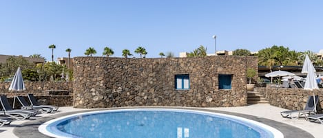A swimming pool with lounge chairs and umbrellas, surrounded by a stone wall and palm trees.
