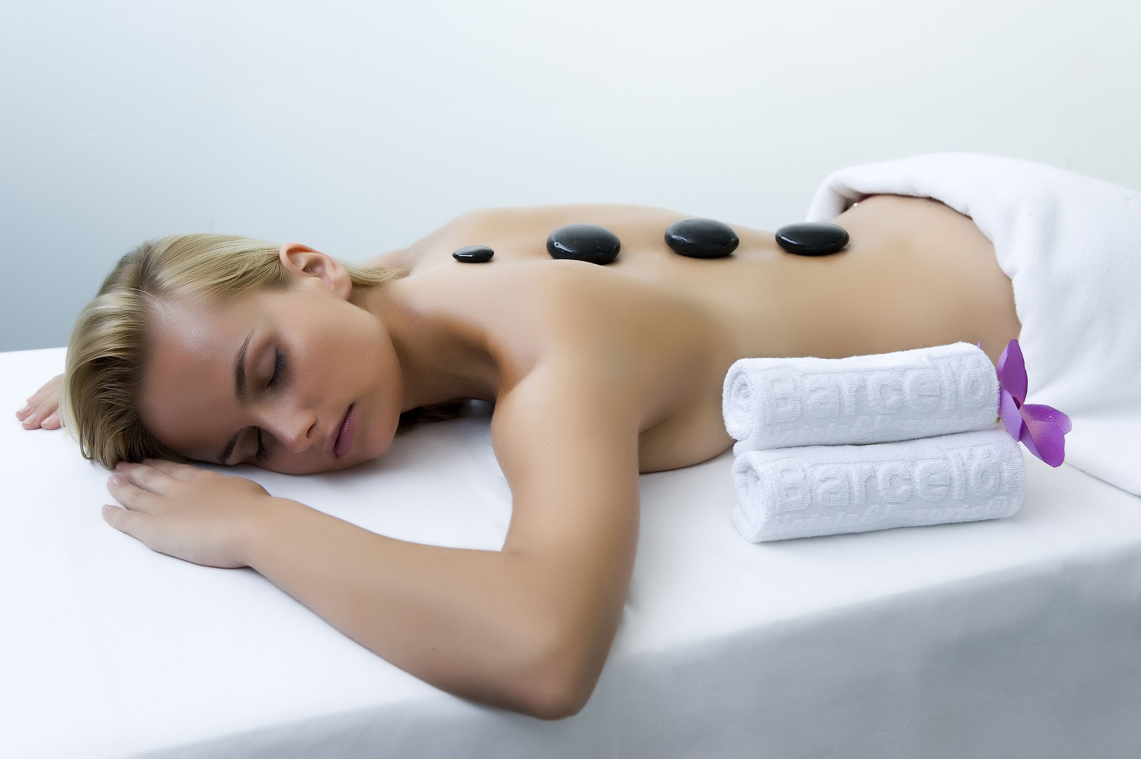 Een persoon ontvangt een rugmassage met stenen op een witte massagetafel, met handdoeken en een paars lint.