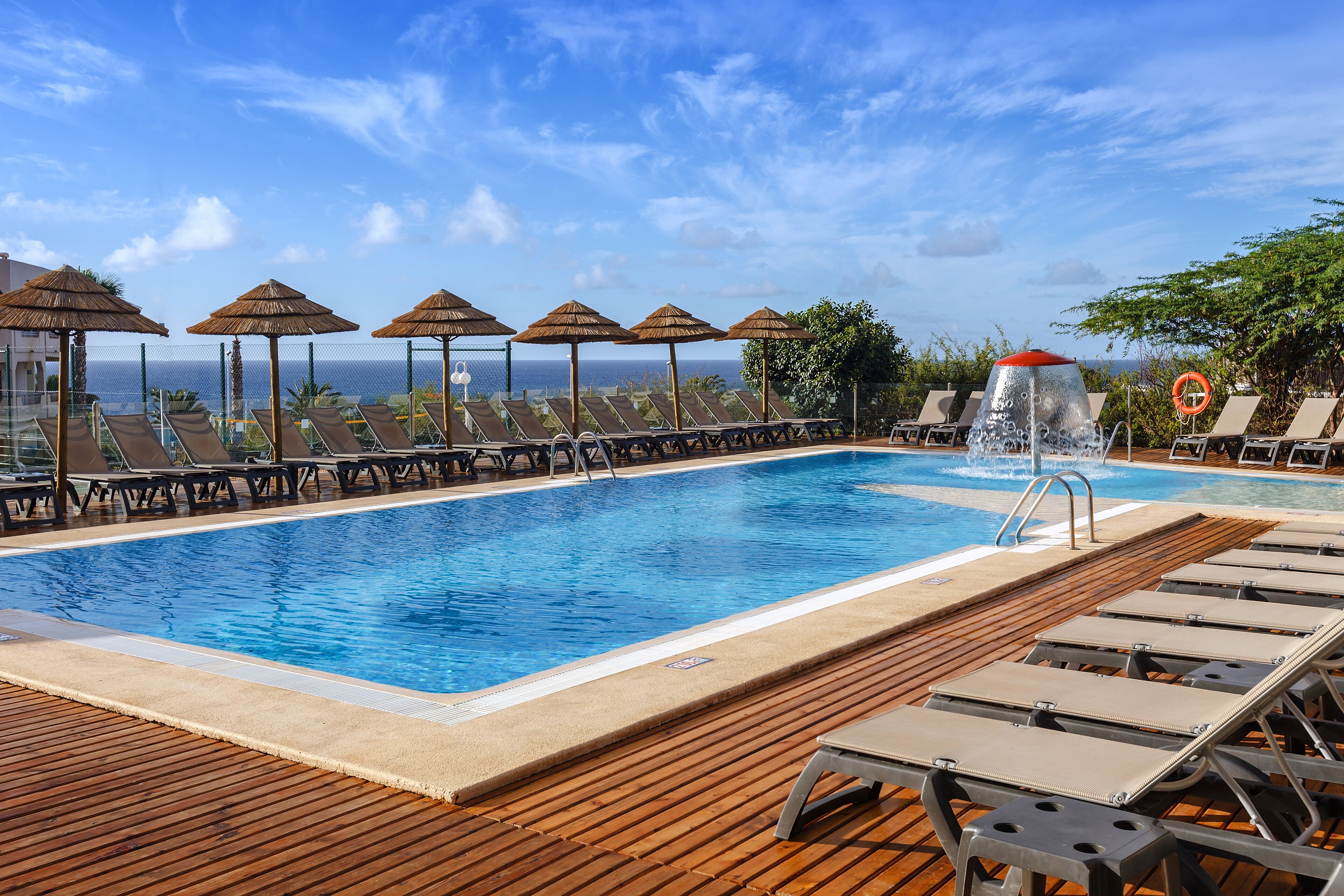 Un área de piscina con sillones de descanso y sombrillas, una terraza de madera y vistas al océano.