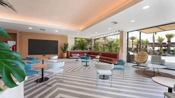A modern indoor lounge area with striped flooring, multiple seating options including sofas and chairs, a large flat-screen TV, and a view of greenery outside.