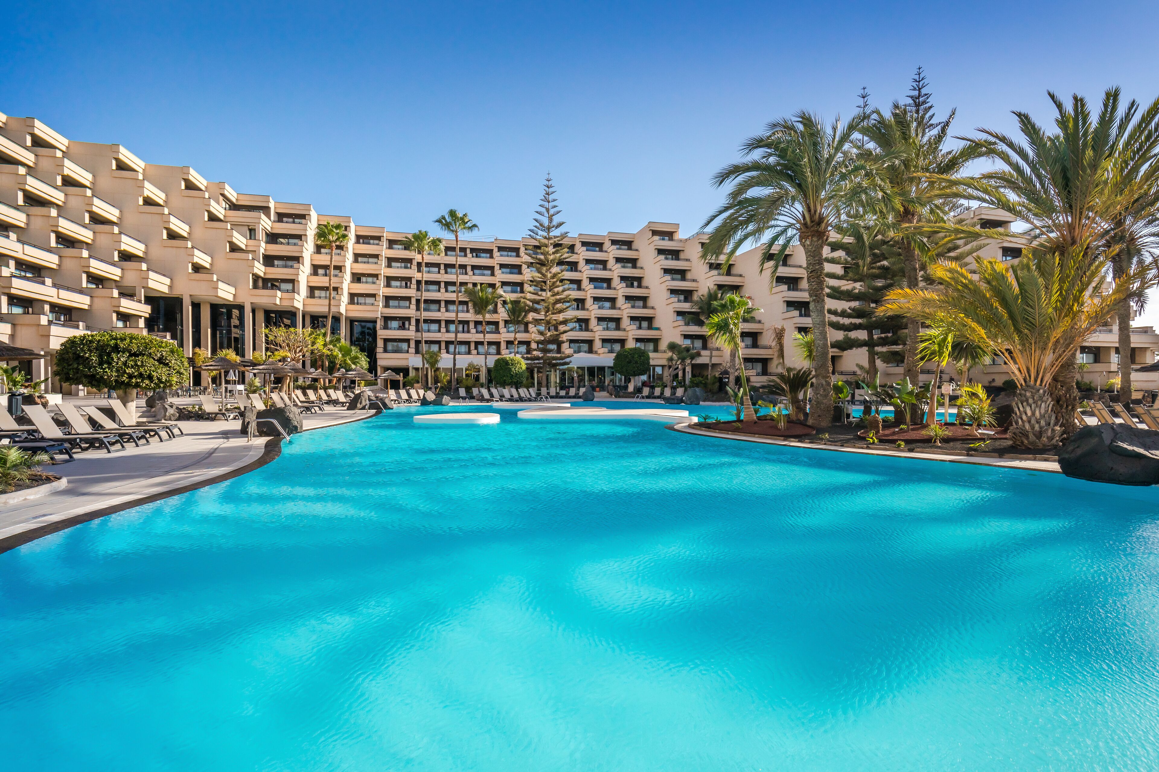 A large swimming pool with lounge chairs and palm trees, surrounded by a multi-story hotel building.