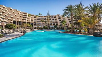 A large swimming pool with lounge chairs and palm trees, surrounded by a multi-story hotel building.