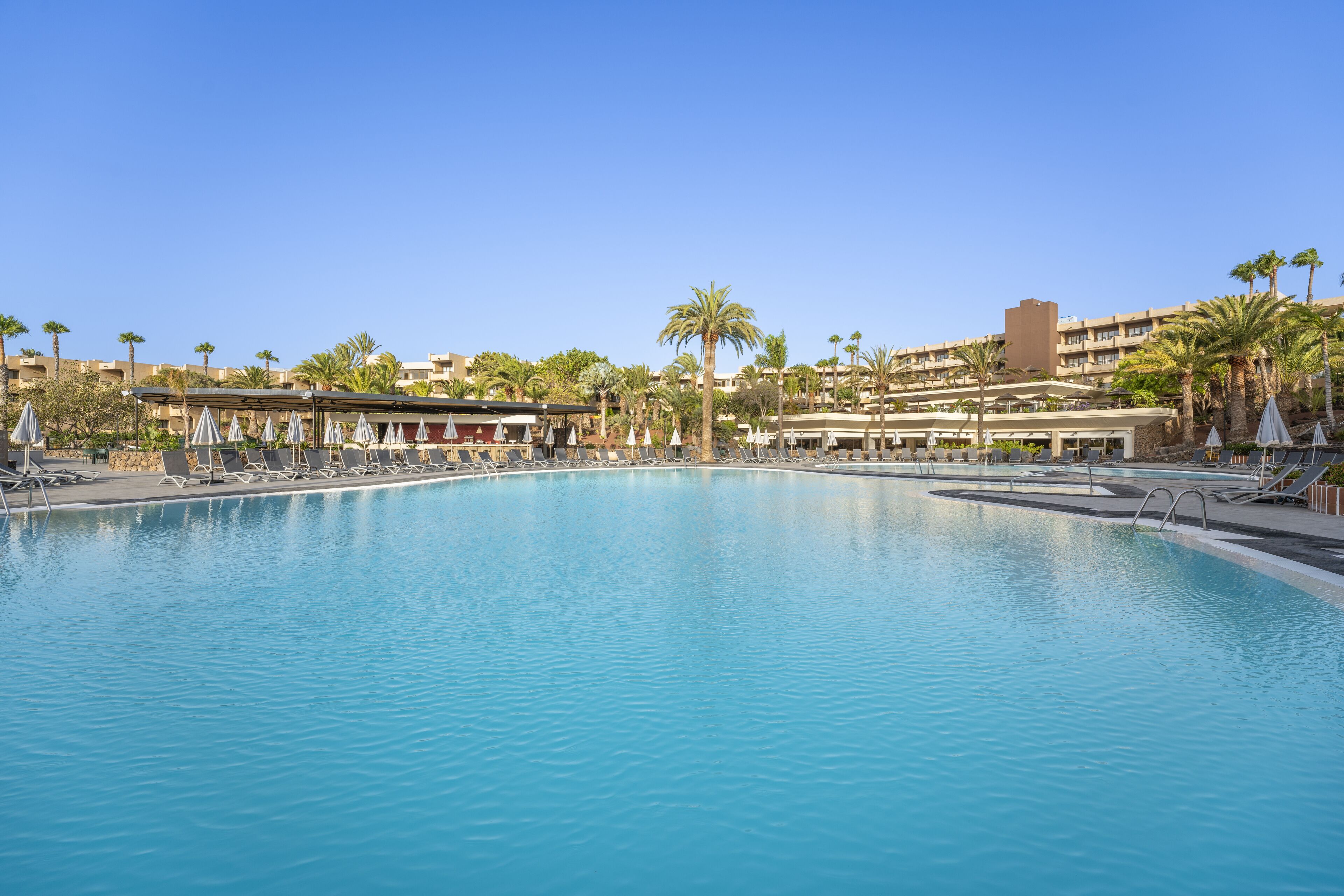 A large, clear swimming pool with lounge chairs and umbrellas, surrounded by palm trees and hotel buildings.