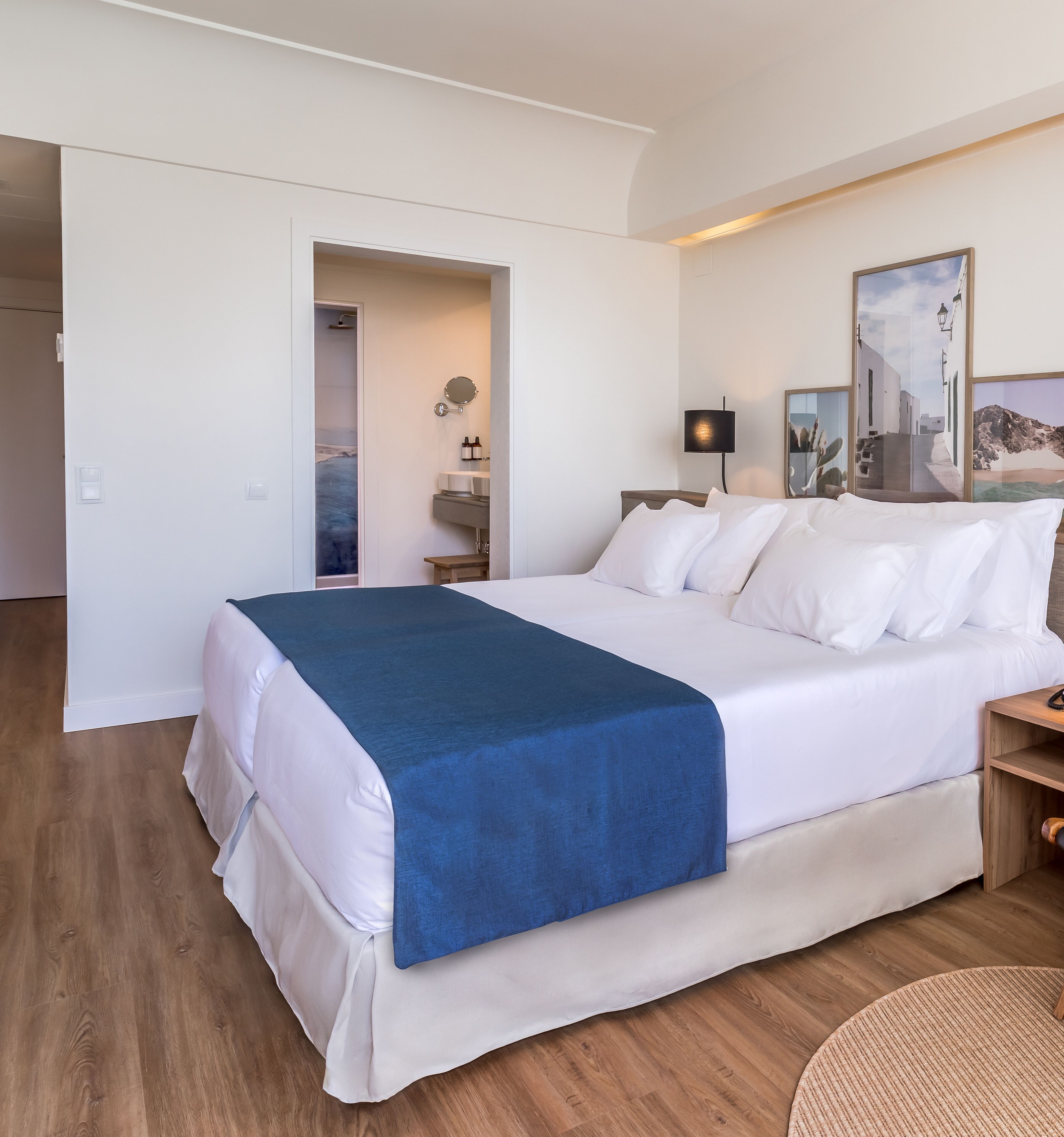 A hotel room with a large bed, a bedside table, a nightstand, a lamp, a mirror, a sink, and a shower.