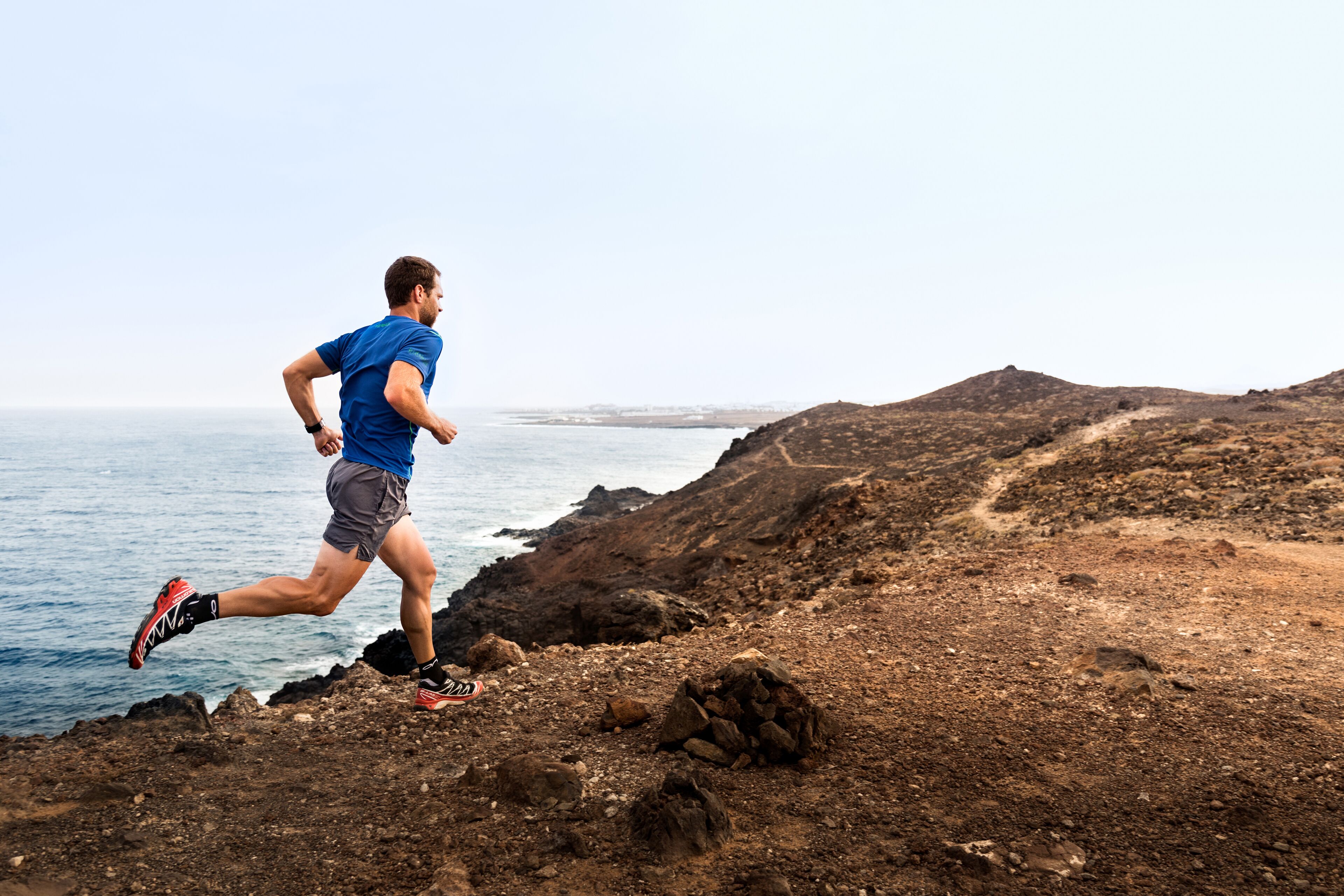 A man running along a rocky coastal path with a view of the sea and distant hills.