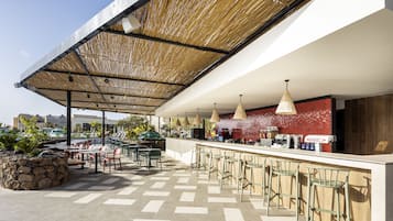 Un área de bar al aire libre con una jardinera de piedra, varias mesas y sillas, y una barra con taburetes.