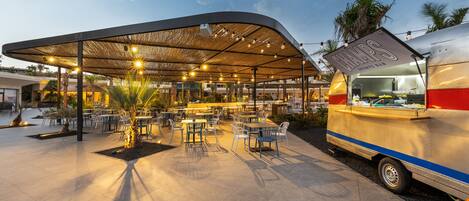 Un área de estar al aire libre con mesas y sillas, un pabellón cubierto y un camión de comida identificado como "CHAMP'S SPORTS BAR".