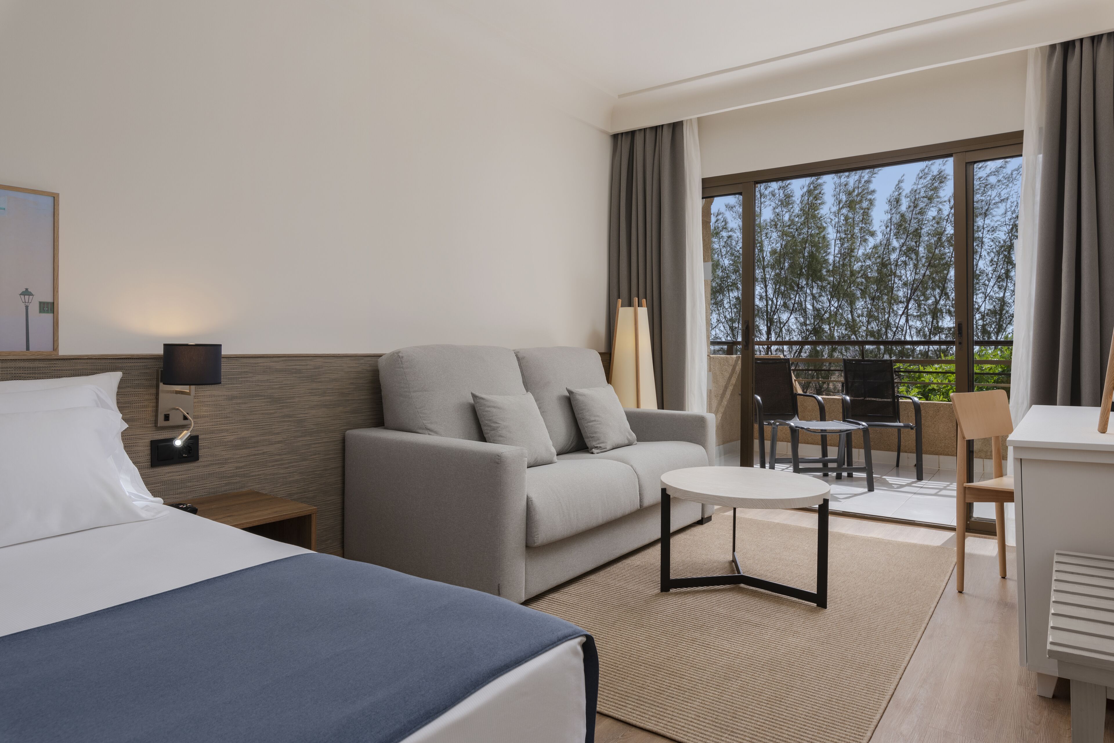 A hotel room with a bed, a sofa, a small table, a desk, and a balcony with chairs.