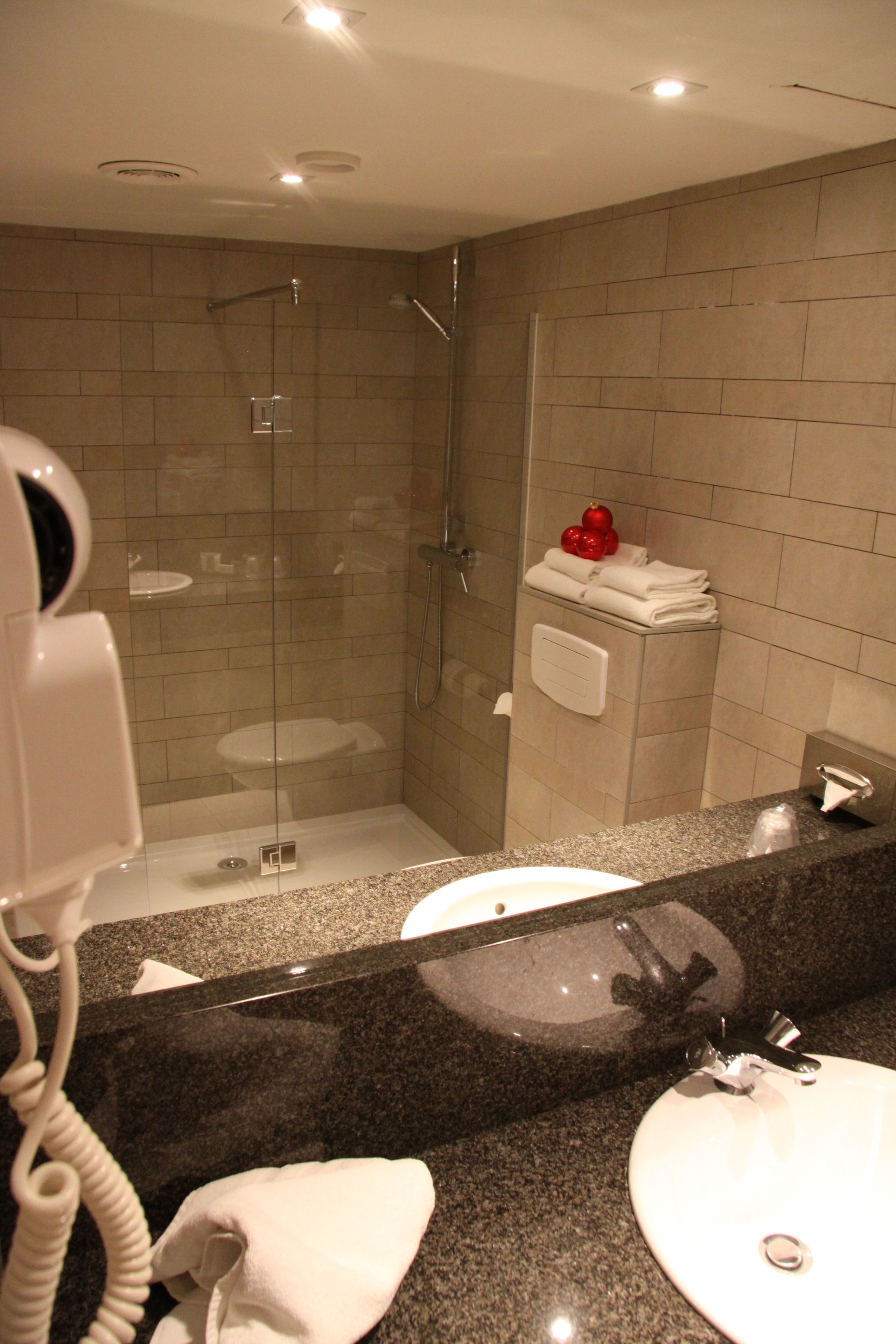 A modern bathroom with a granite countertop, two sinks, a glass-enclosed shower, and a hairdryer on the wall.