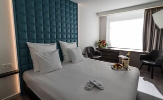 Habitación de hotel con una cama grande, un cabecero azul, dos sillas, una mesita con frutero y una botella, y una ventana con cortinas.