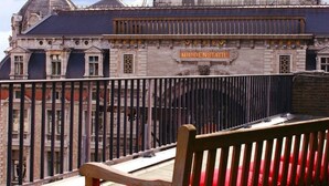 Un banco con un cojín rojo en un balcón adoquinado con vistas a un edificio histórico con una cúpula y el letrero "MIDDENSTATIE".