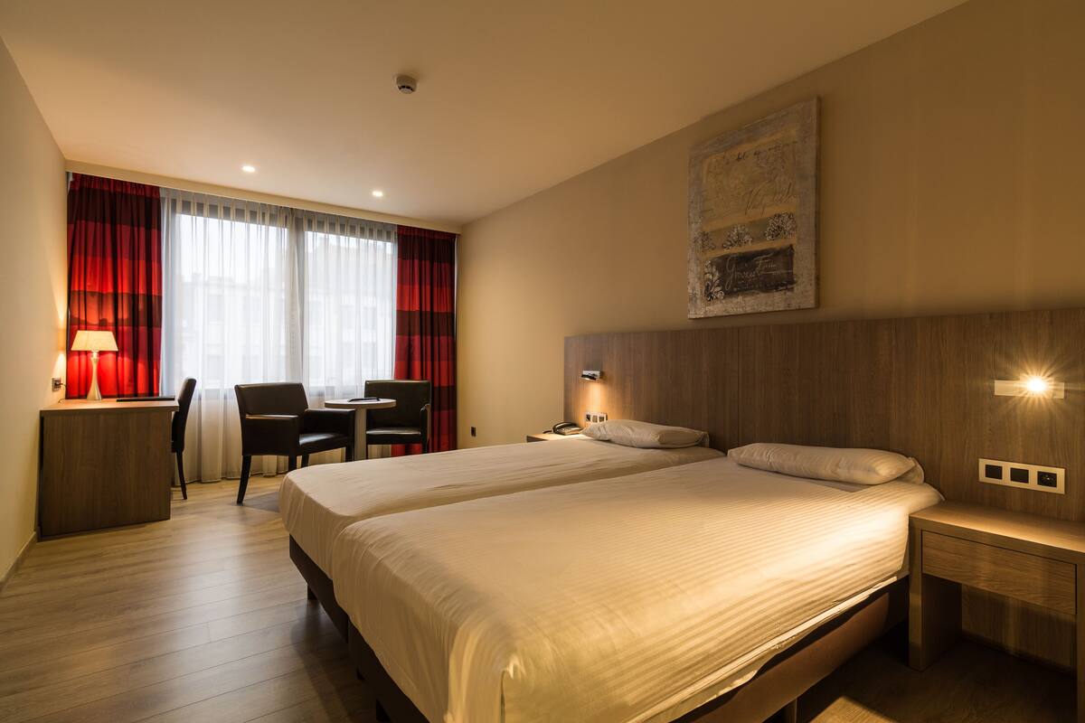 Habitación de hotel con dos camas, un escritorio con silla, una lámpara y un ventanal con cortinas rojas.