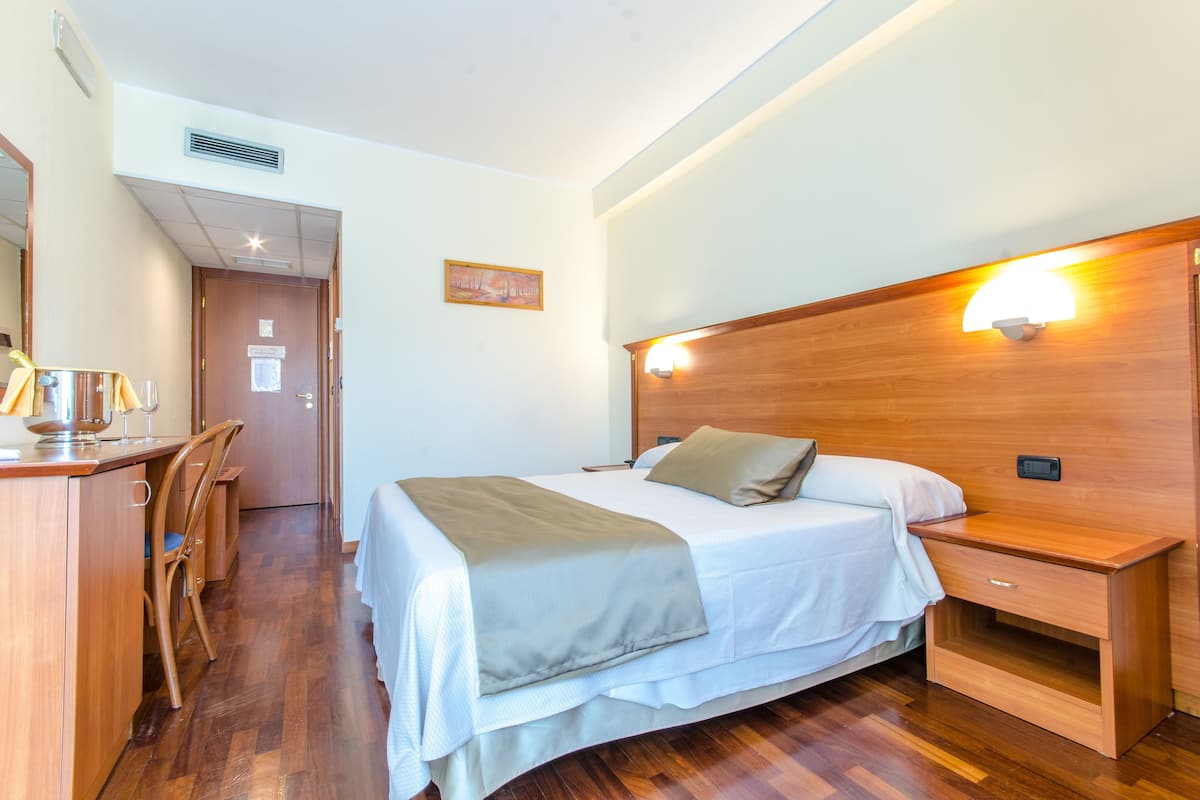 A hotel room with a wooden bed, bedside table, and a desk with chairs.
