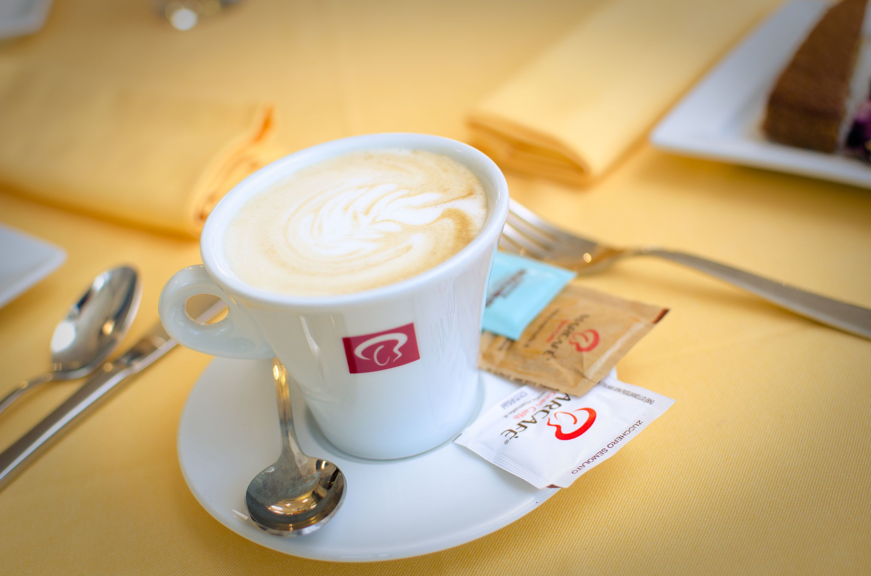 Una taza de café con arte de latte sobre un plato, una cuchara, un sobre de azúcar y una servilleta sobre un mantel amarillo.