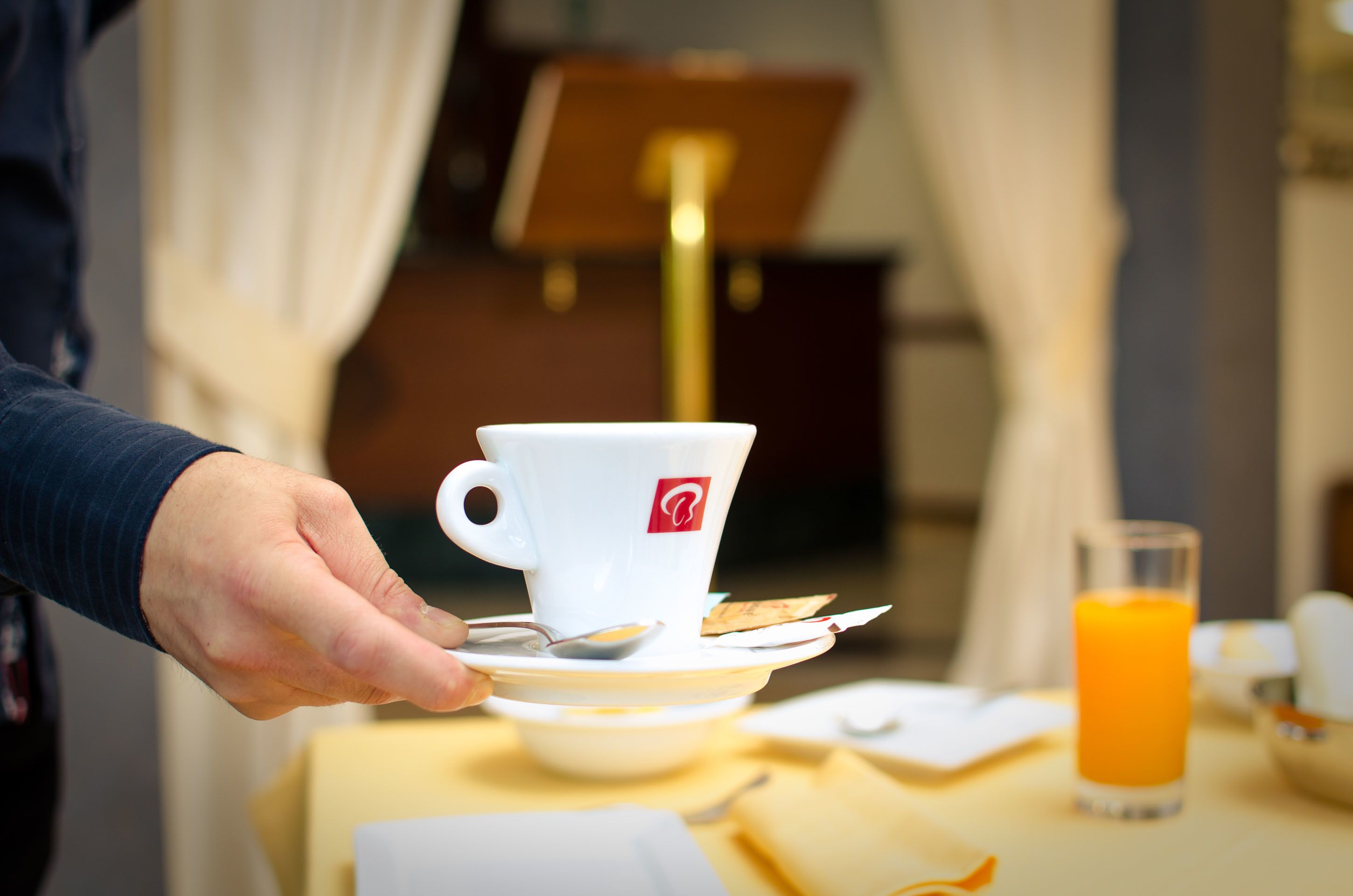 A hand holding a white cup with a logo, a glass of orange juice, and a spoon on a saucer.