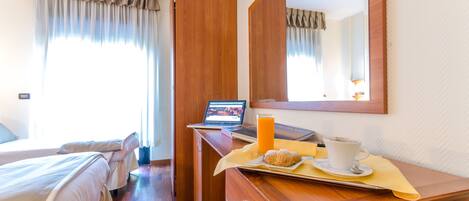 Quarto de hotel com uma cama, uma escrivaninha com um laptop, uma chávena de café e um tabuleiro com um pastel.