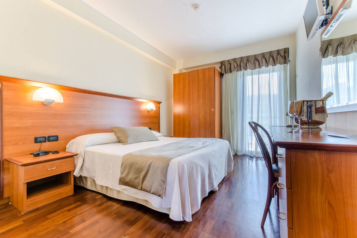 A hotel room with a wooden bed, bedside table, lamp, and a desk with a chair.