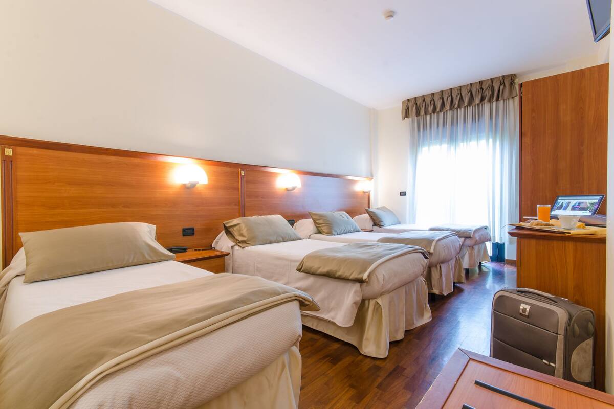 A hotel room with three beds, wooden headboards, and a desk with a computer and a cup of coffee.