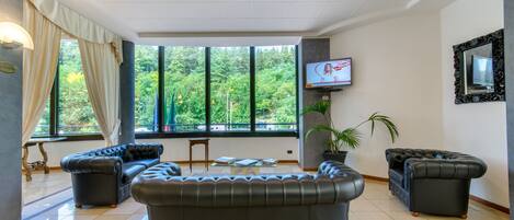 A spacious living room with a large sectional sofa, a television mounted on the wall, and a view of lush greenery through large windows.
