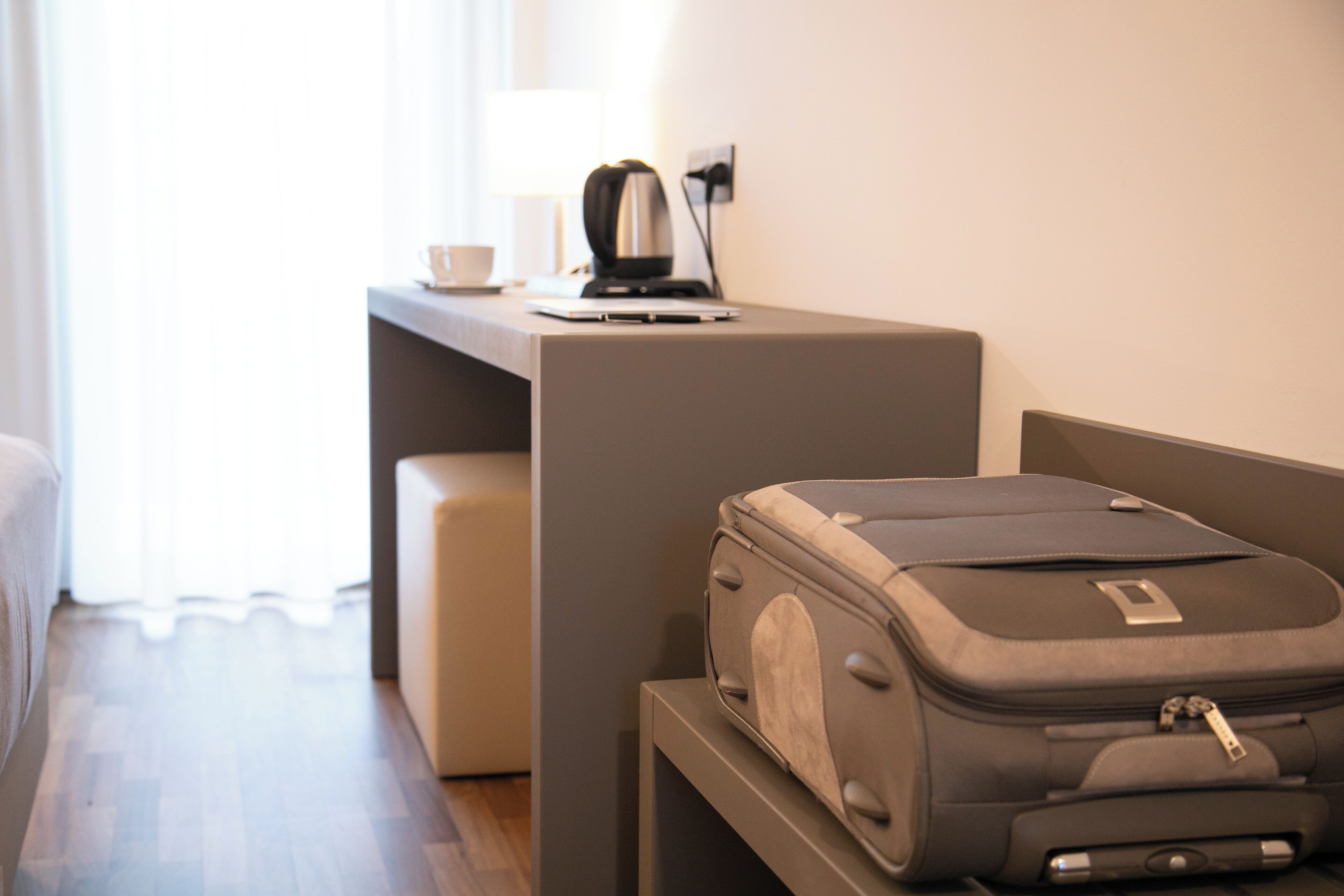 Una habitación de hotel con cama, escritorio, laptop, tetera y maleta.