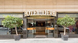 A hotel entrance with a modern facade, two potted trees, and a glass door.