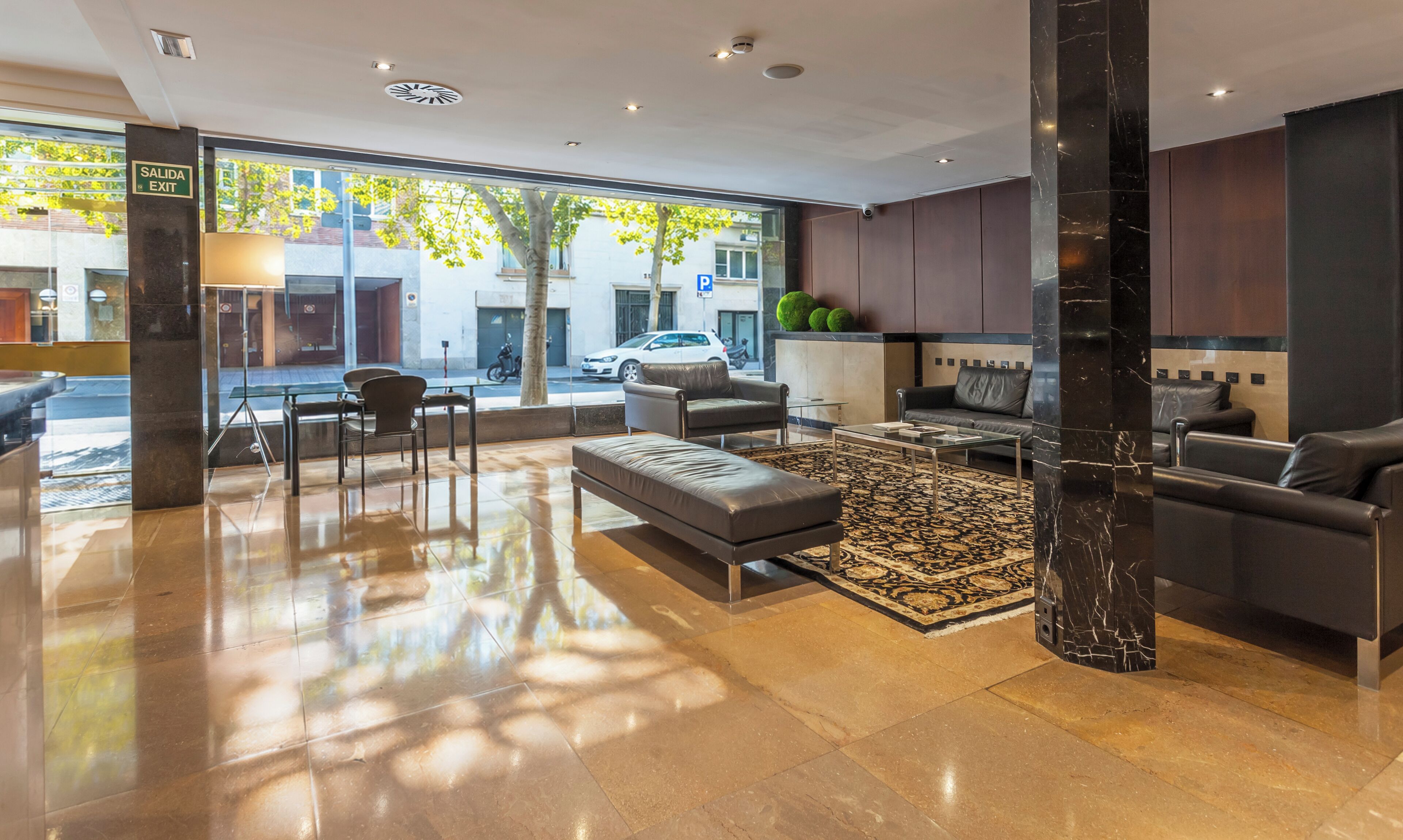 A modern lobby with a glass exit door, a seating area with a sofa and armchairs, a coffee table, and a patterned rug.