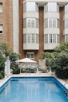 A swimming pool with a white umbrella, surrounded by greenery and a brick building with multiple windows.