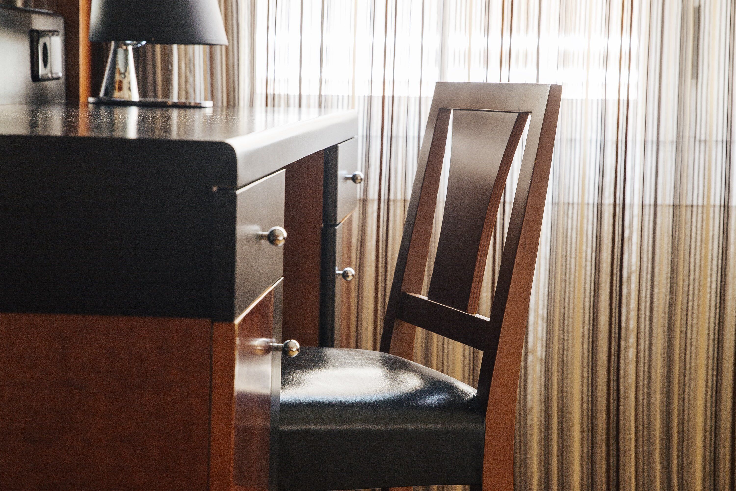 Een houten bureau met een lamp, een stoel en een raam met verticale jaloezieën.