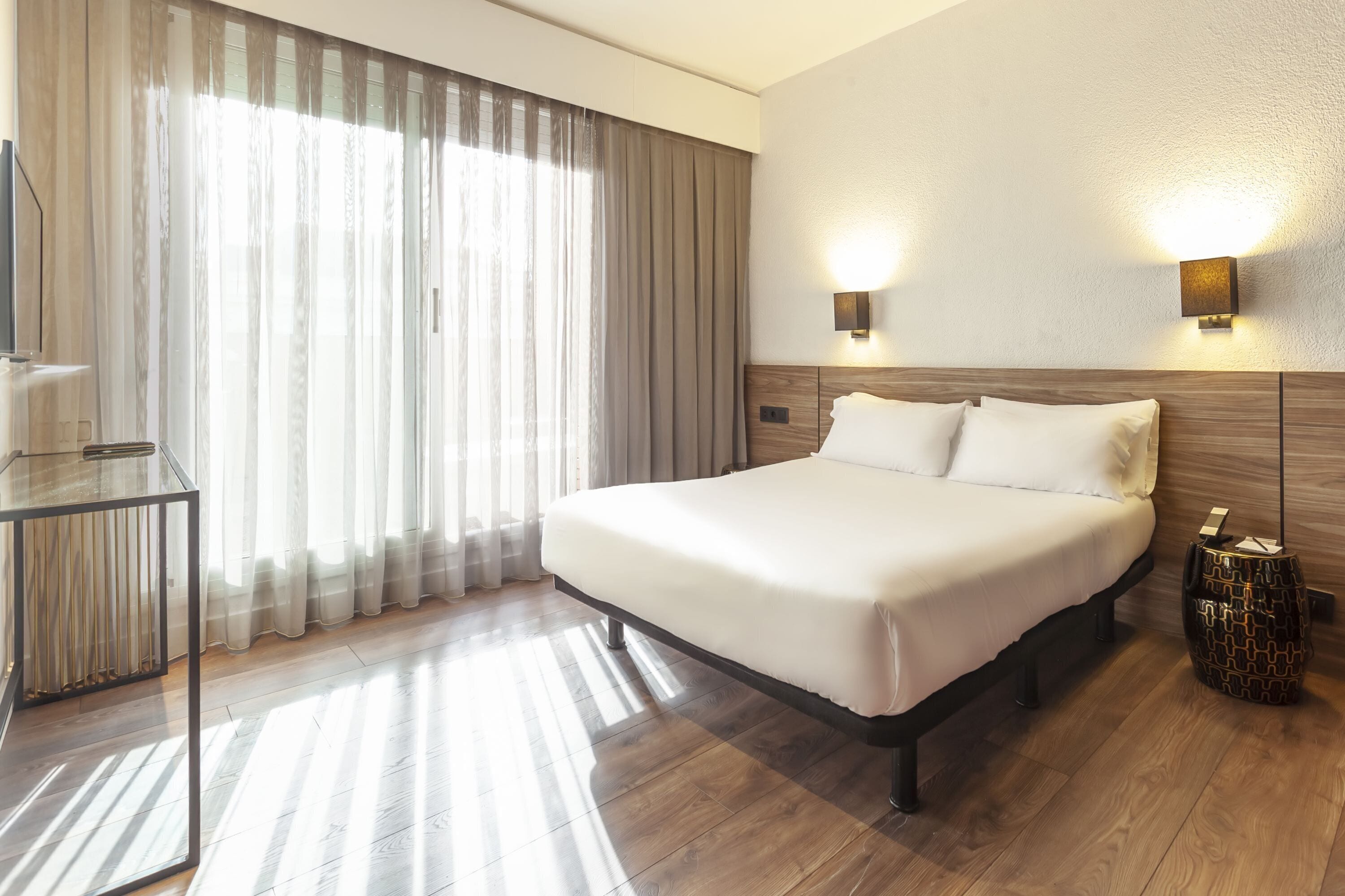 Una habitación de hotel con una cama grande, una mesita de noche, un televisor y una ventana con cortinas.