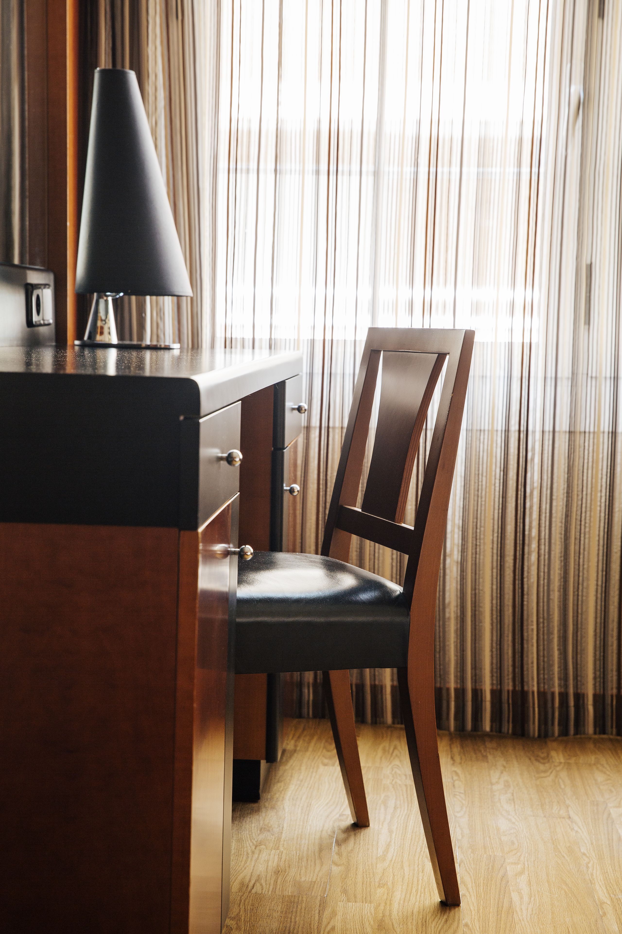 A wooden desk with a lamp and a chair in front of a window with curtains.