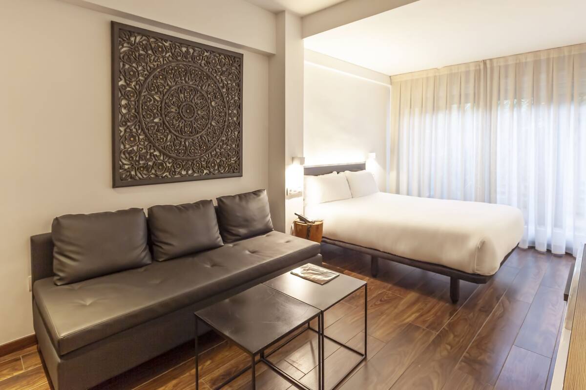 A modern hotel room with a sofa, a bed, a small table, and a decorative wall piece.