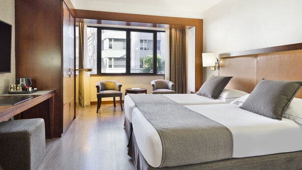 A hotel room with two beds, a desk, a chair, a TV, and a window with a view of buildings.