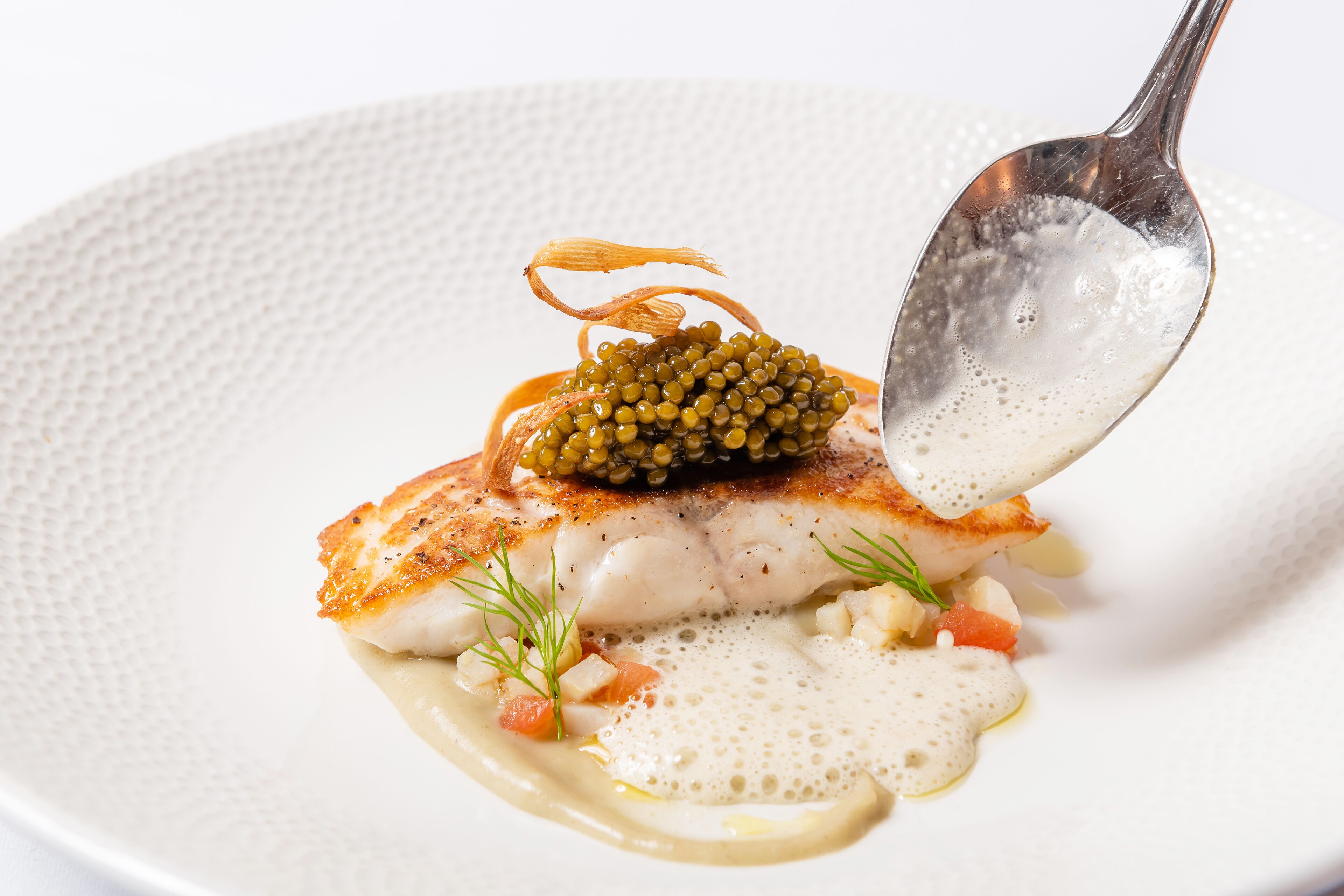 A white plate with a piece of fish topped with caviar, garnished with dill and a sauce.