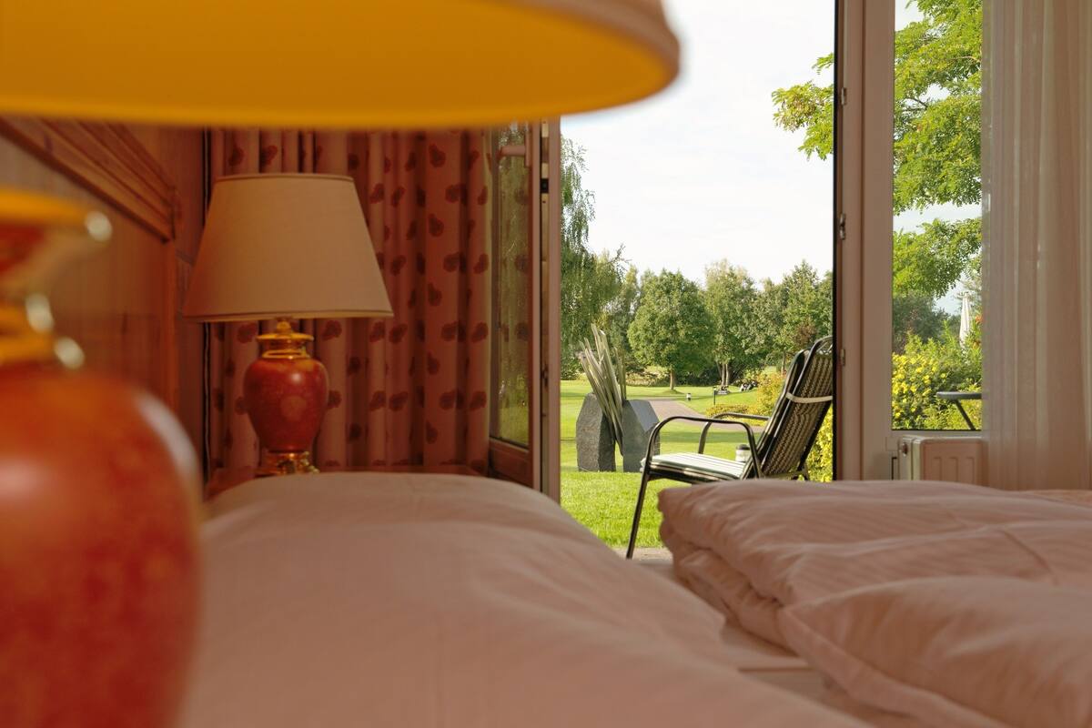 A hotel room with a bed, a lamp, and a view of a garden and outdoor seating area.