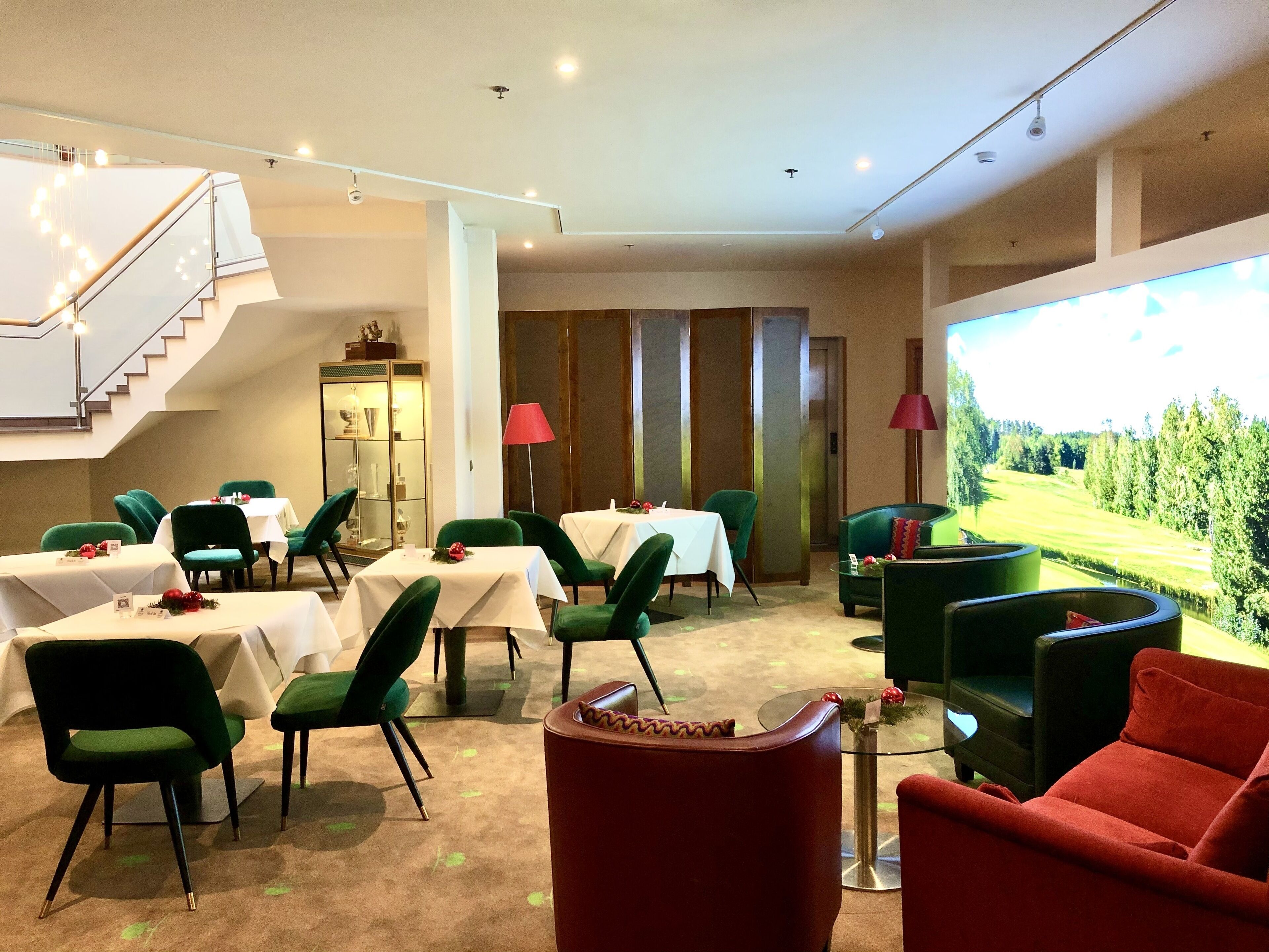 A hotel dining area with tables and chairs, a staircase, a cabinet, and a large window with a view of a green landscape.
