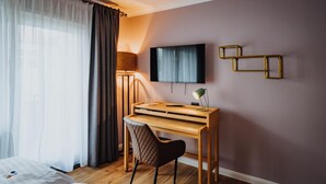 Quarto de hotel com uma escrivaninha de madeira, uma televisão fixada na parede e uma cama com roupa de cama branca.