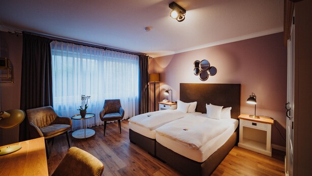 Quarto de hotel com duas camas, uma escrivaninha, uma cadeira, um abajur e uma mesa de cabeceira.