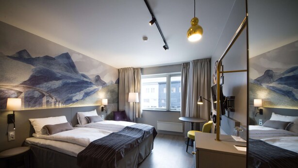 A hotel room with two beds, a desk, a chair, a mirror, and a window with curtains.