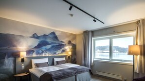 A hotel room with a large bed, two bedside tables, a window with a view of the sea, and a painting on the wall.