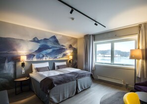 A hotel room with a large bed, two bedside tables, a window with a view of the sea, and a painting on the wall.