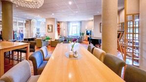 A spacious hotel lobby with a long wooden table, comfortable seating, a large chandelier, and a view of the outdoor area through large windows. - Golfhotel Fahrenbach (Trostau)