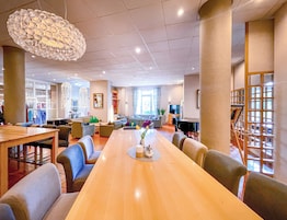 Um hall de hotel espaçoso com uma longa mesa de madeira, assentos confortáveis, um grande lustre e vista para a área externa por grandes janelas.