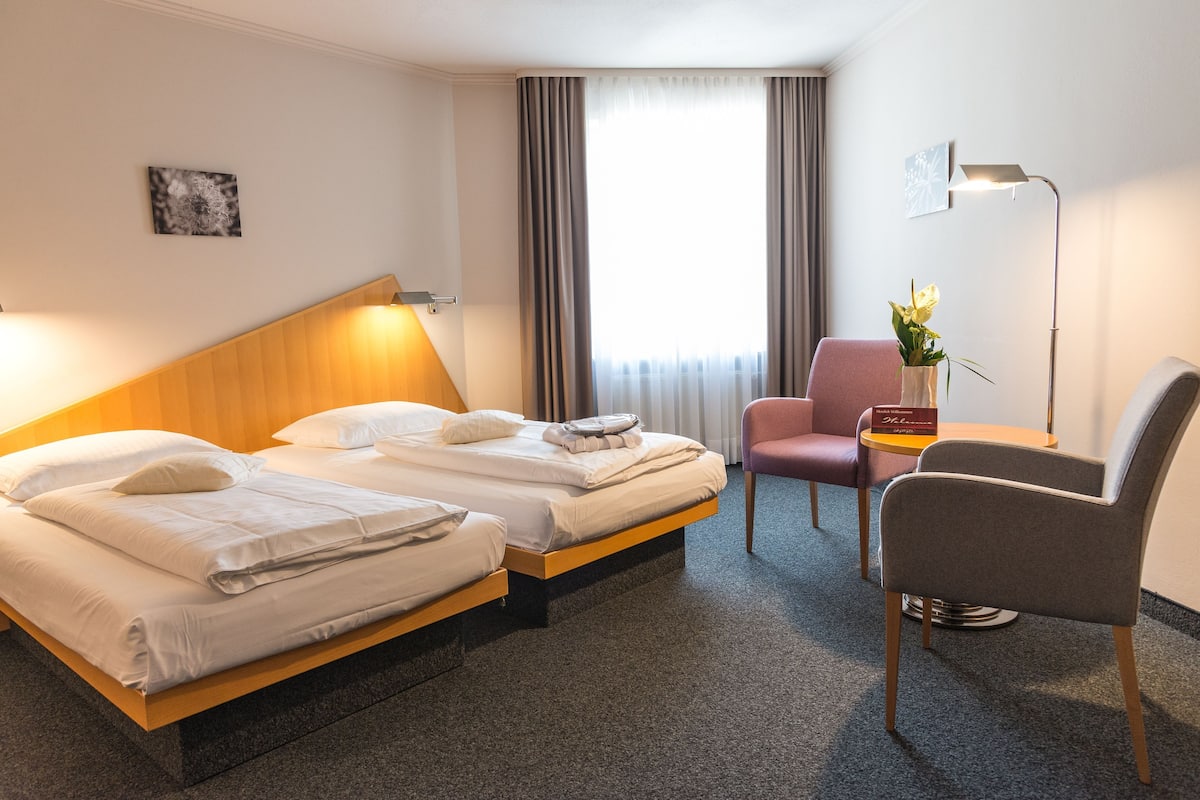 A hotel room with two beds, a desk, a chair, a lamp, and a window with curtains.