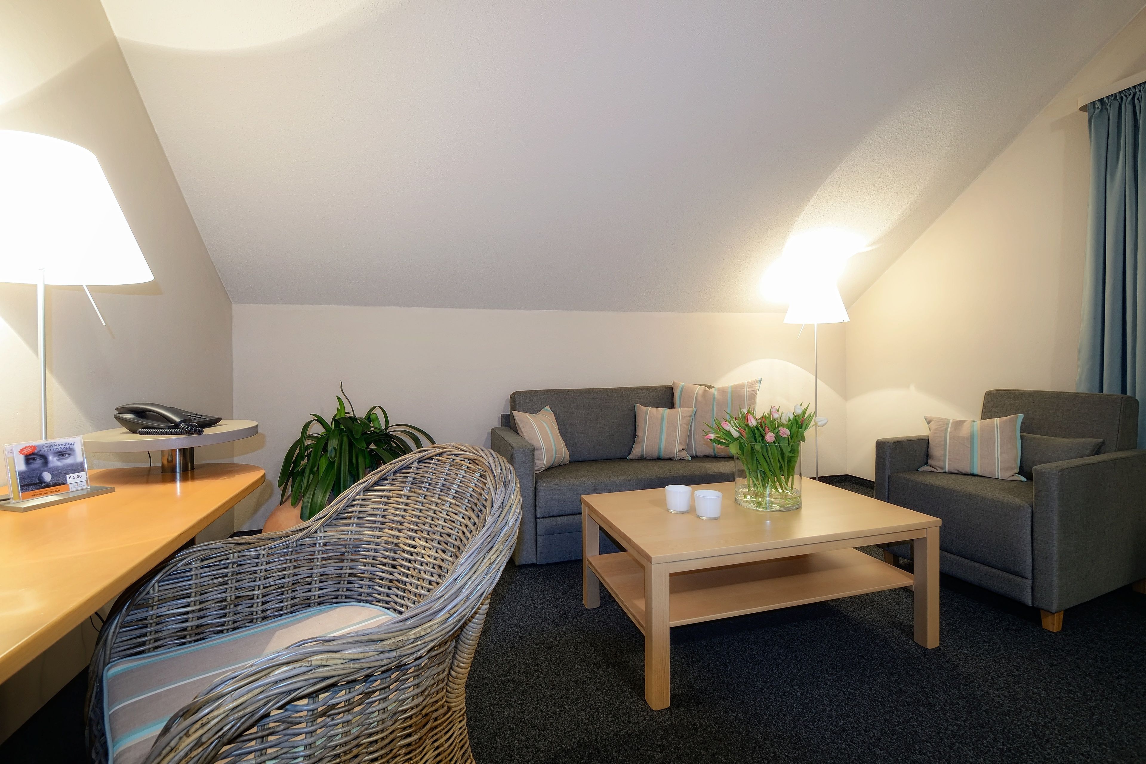 Un salon moderne avec un canapé, un fauteuil, une table basse et une chaise en osier. Il y a une lampe sur le bureau et une plante sur le sol.